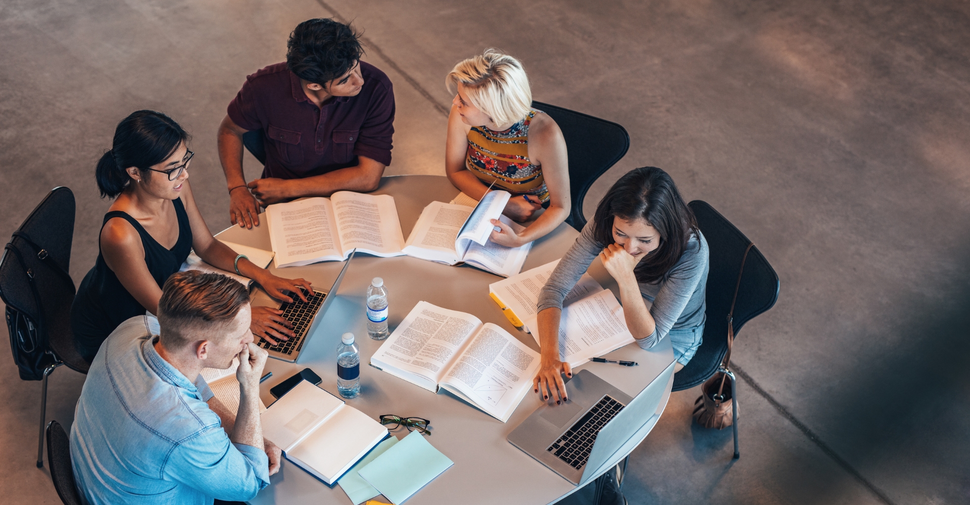 Five students sit around a round table with textbooks and laptops, studying together.